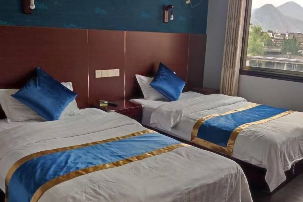 A hotel room with two beds, each with a blue and gold bedspread, a wooden headboard, a nightstand, and a window with a view of mountains.