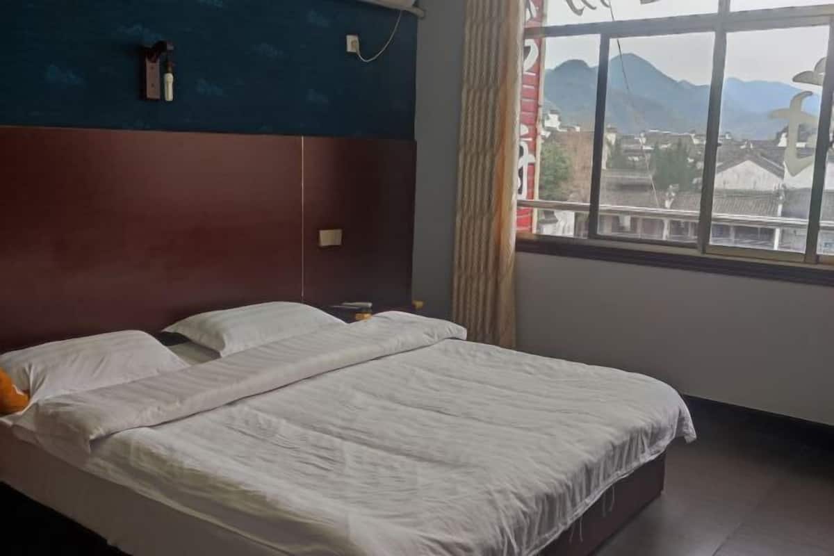 A double bed with white bedding, a wooden headboard, a nightstand with a lamp, and a window with a view of buildings and mountains.