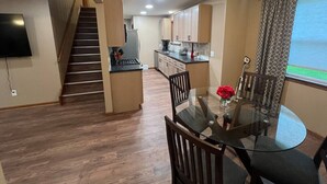 A modern kitchen with wooden cabinets, a dining area with a glass table, and a staircase leading to another level.