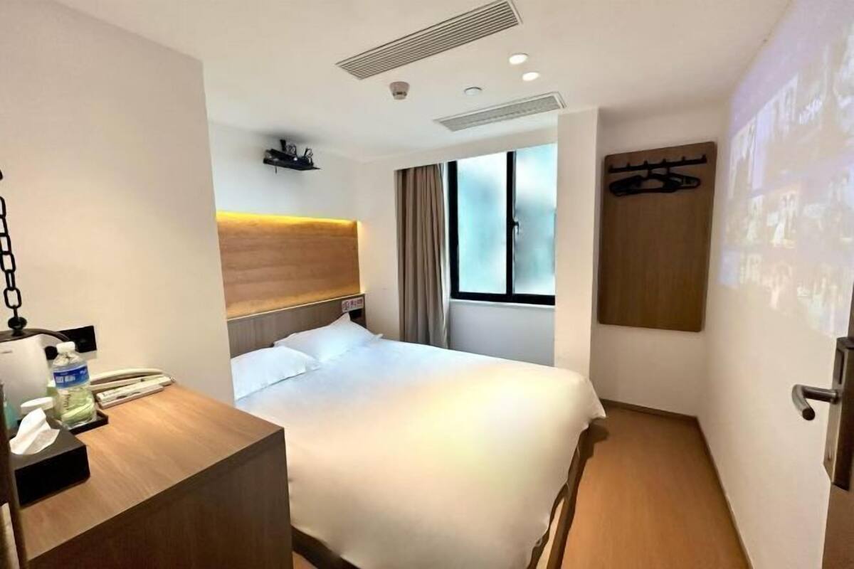 Ein Hotelzimmer mit einem Bett, einem Schreibtisch mit Telefon und Flasche, einem Kopfteil, einem Fenster mit Vorhängen und einer Tür mit Kleiderhaken.