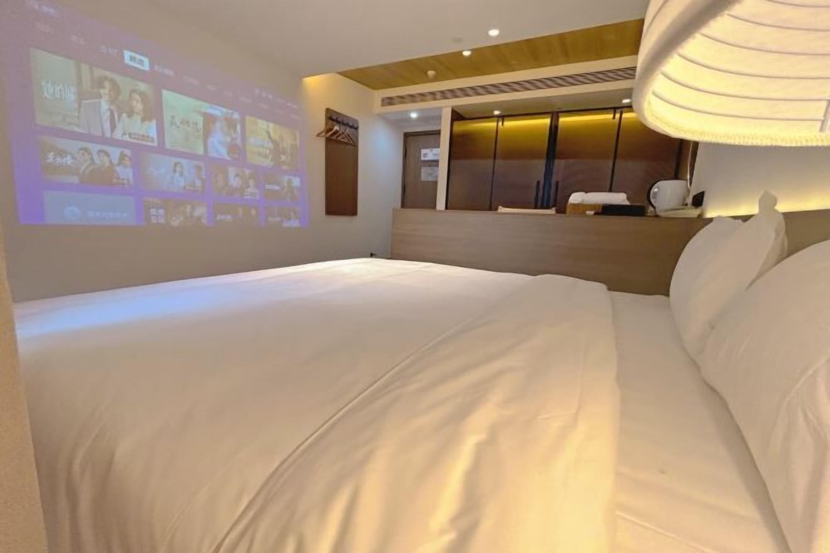 A hotel room with a bed, a wall-mounted TV displaying a movie, a wardrobe, and a ceiling light.