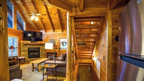 A cozy cabin interior with a stone fireplace, wooden walls, a television, and a refrigerator. - Family Cabin with Hot Tub, Game Room & Pool Access (Pigeon Forge)