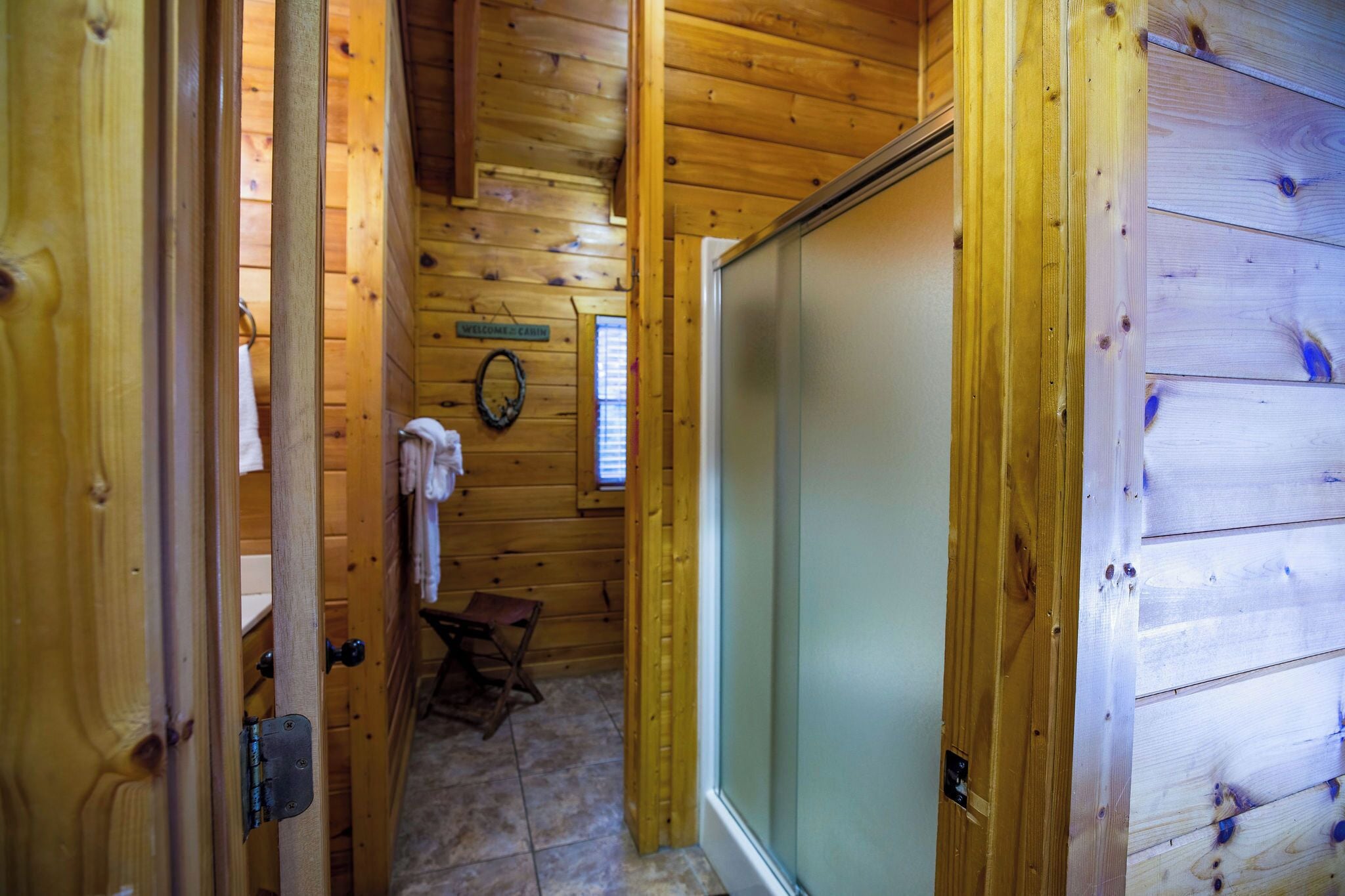 A wooden cabin interior with a bathroom featuring a shower, a towel rack, and a small wooden stool.