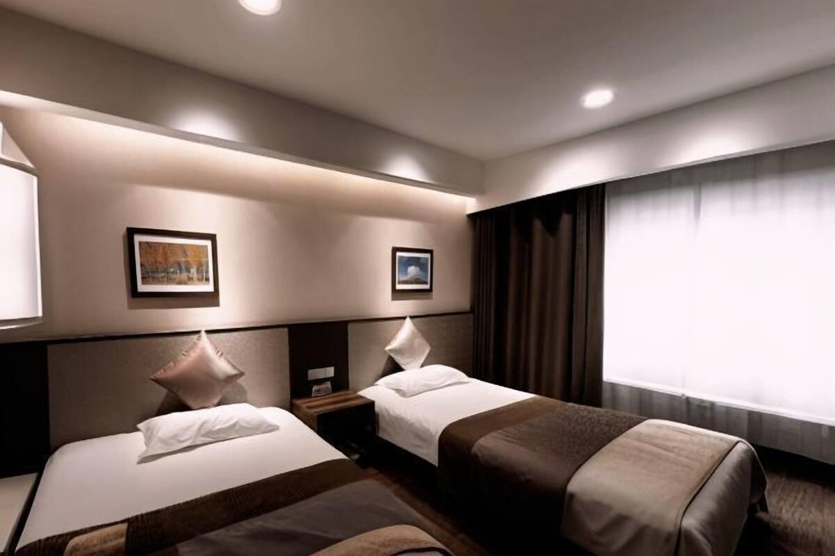 A hotel room with two beds, a nightstand, a window with curtains, and two framed pictures on the wall.
