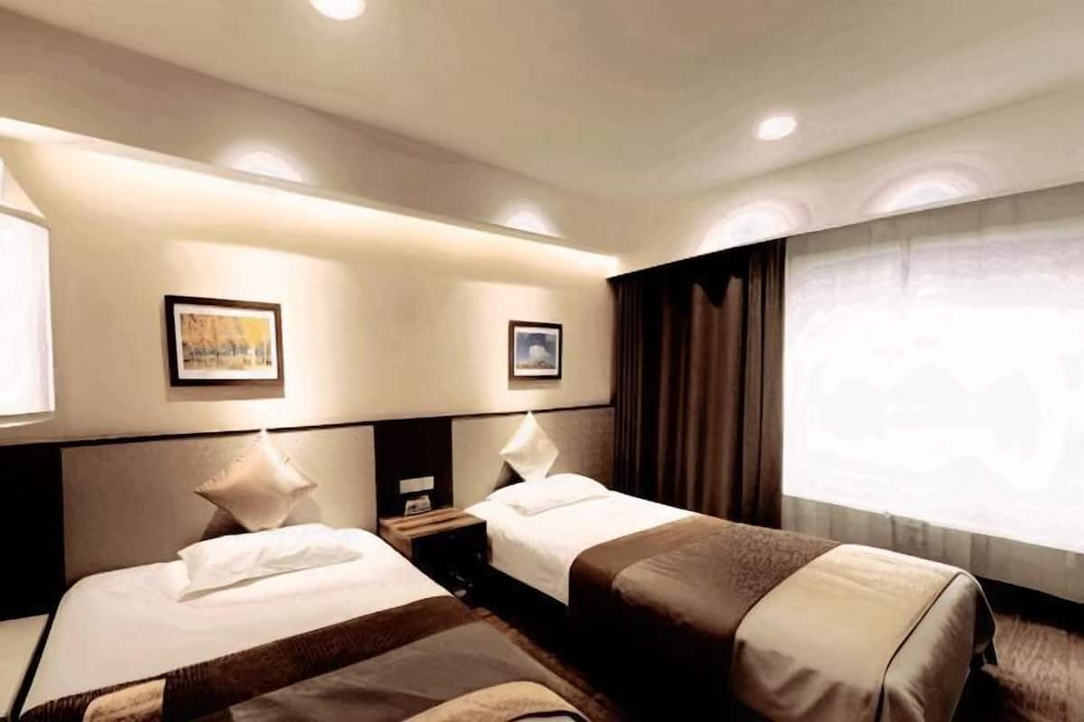 A hotel room with two beds, a nightstand, a wall-mounted lamp, and a window with curtains.