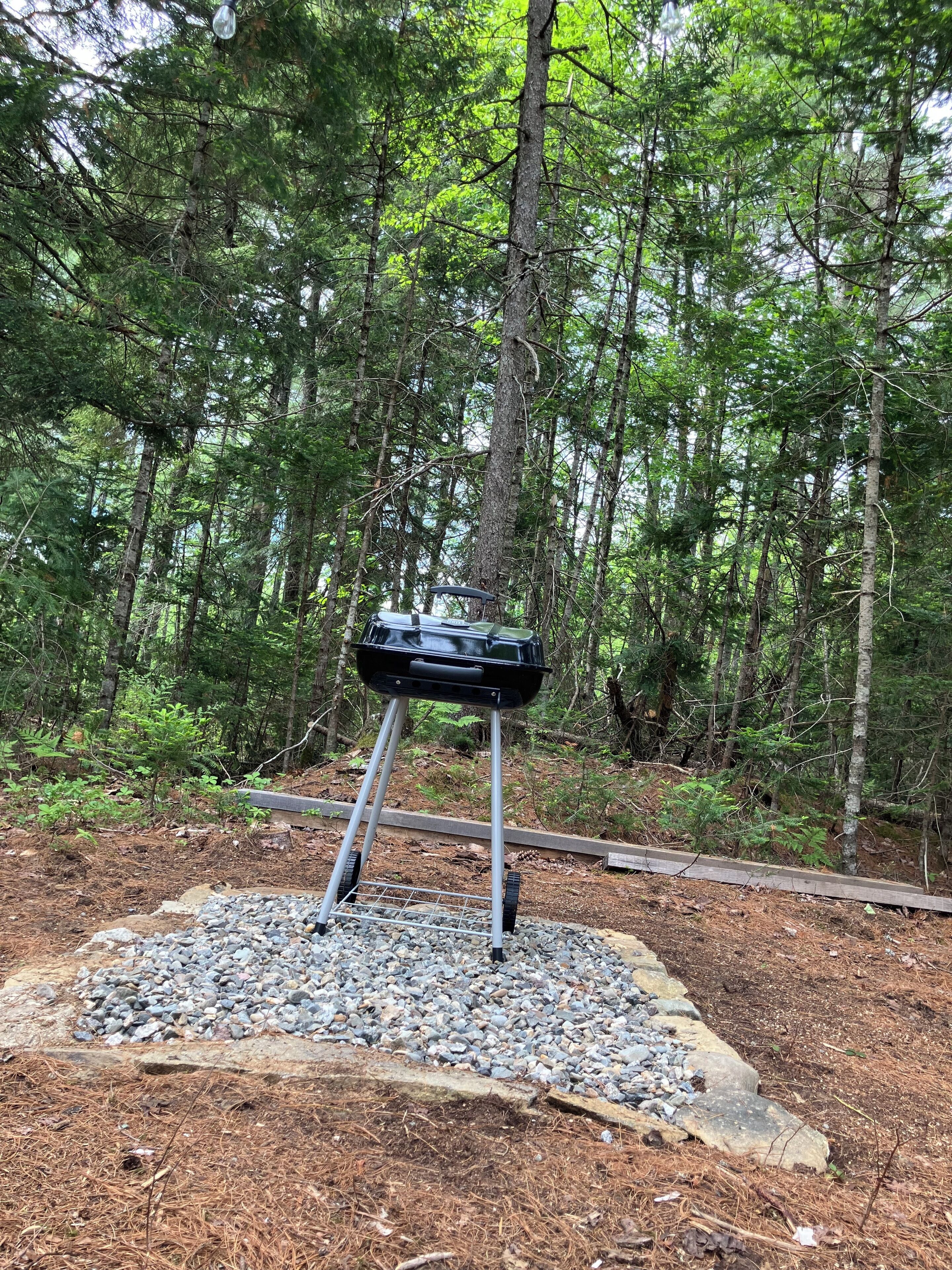 A portable grill set up on a gravel platform surrounded by a forest.