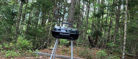 A portable grill set up on a gravel platform surrounded by a forest.
