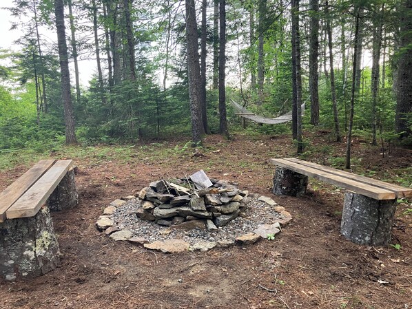 Un claro en el bosque con un hoyo para el fuego rodeado de rocas y troncos, dos bancos de madera y una hamaca.