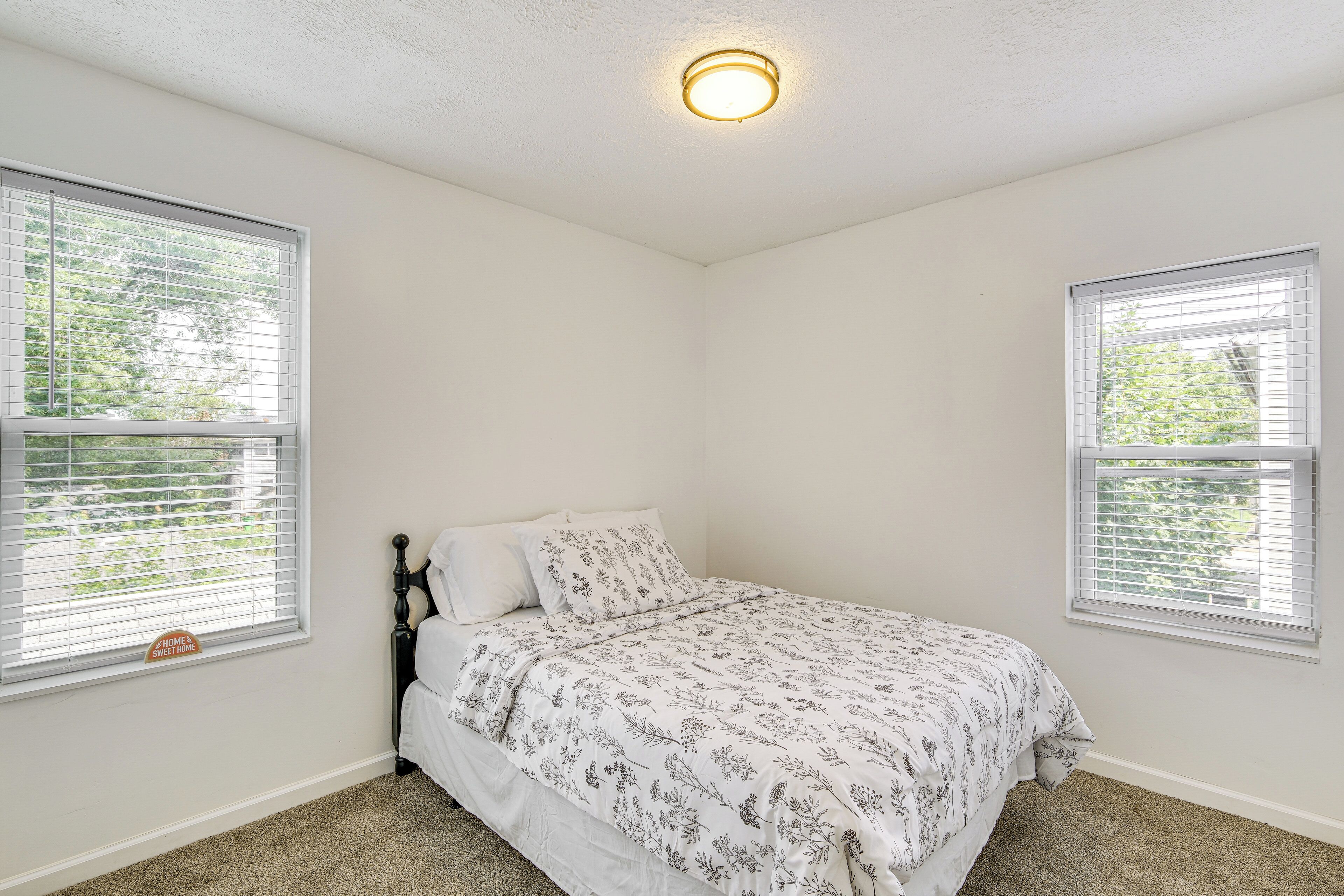 A bedroom with a bed, two windows with blinds, a ceiling light, and a carpeted floor.