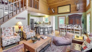 A spacious living area with a wooden staircase, a kitchen with white cabinets, and a dining area with bar stools. - Peaceful Greers Ferry Cabin w/ Deck & Lake View! (Greers Ferry)