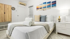 A neatly made bed with white linens and a rolled towel, a bedside table with a bottle and glasses, and a wall with three landscape paintings. - Cozy 3BR Town Home in Gated Community @ Palm Beach (Noord)