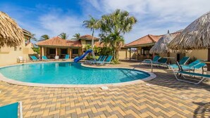 A swimming pool with lounge chairs and a slide, surrounded by palm trees and buildings with thatched roofs. - Cozy 3BR Town Home in Gated Community @ Palm Beach (Noord)