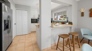 Een moderne keuken met witte kasten, een inbouwoven en een groot raam. In de eethoek staan twee houten krukken en een lichtblauwe stoel.