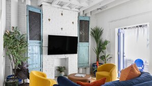 Een woonkamer met een witte bakstenen muur, een flatscreen televisie boven een open haard, gele en blauwe meubels, en potplanten.