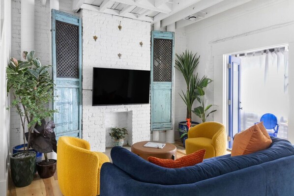 A living room with a white brick wall, a flat-screen TV mounted above a fireplace, yellow and blue furniture, and potted plants. - ★ HostWise Stays ★ Casa Santorini - Aegean Inspired, Instaworthy! (Pittsburgh)