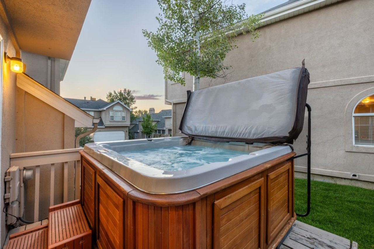 A hot tub with a wooden exterior and a cover, located on a wooden deck with a view of a house and a tree in the background.