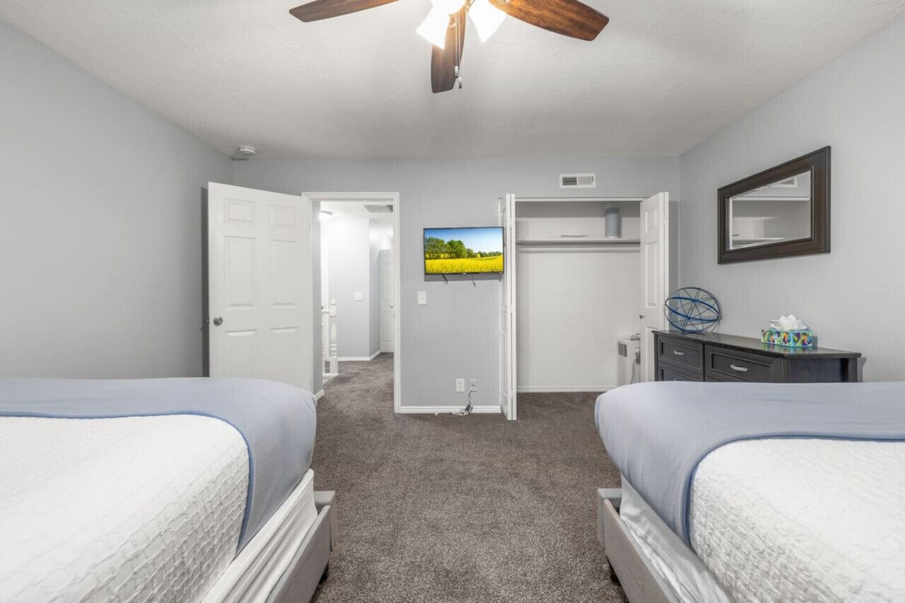 A bedroom with two beds, a ceiling fan, a TV, a wardrobe, and a door leading to another room.