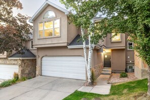 A two-story house with a stone foundation, a white garage door, and a small front yard with a tree. - Oak Ridge Retreat | Hot Tub | Grill | Spacious (Cottonwood Heights)