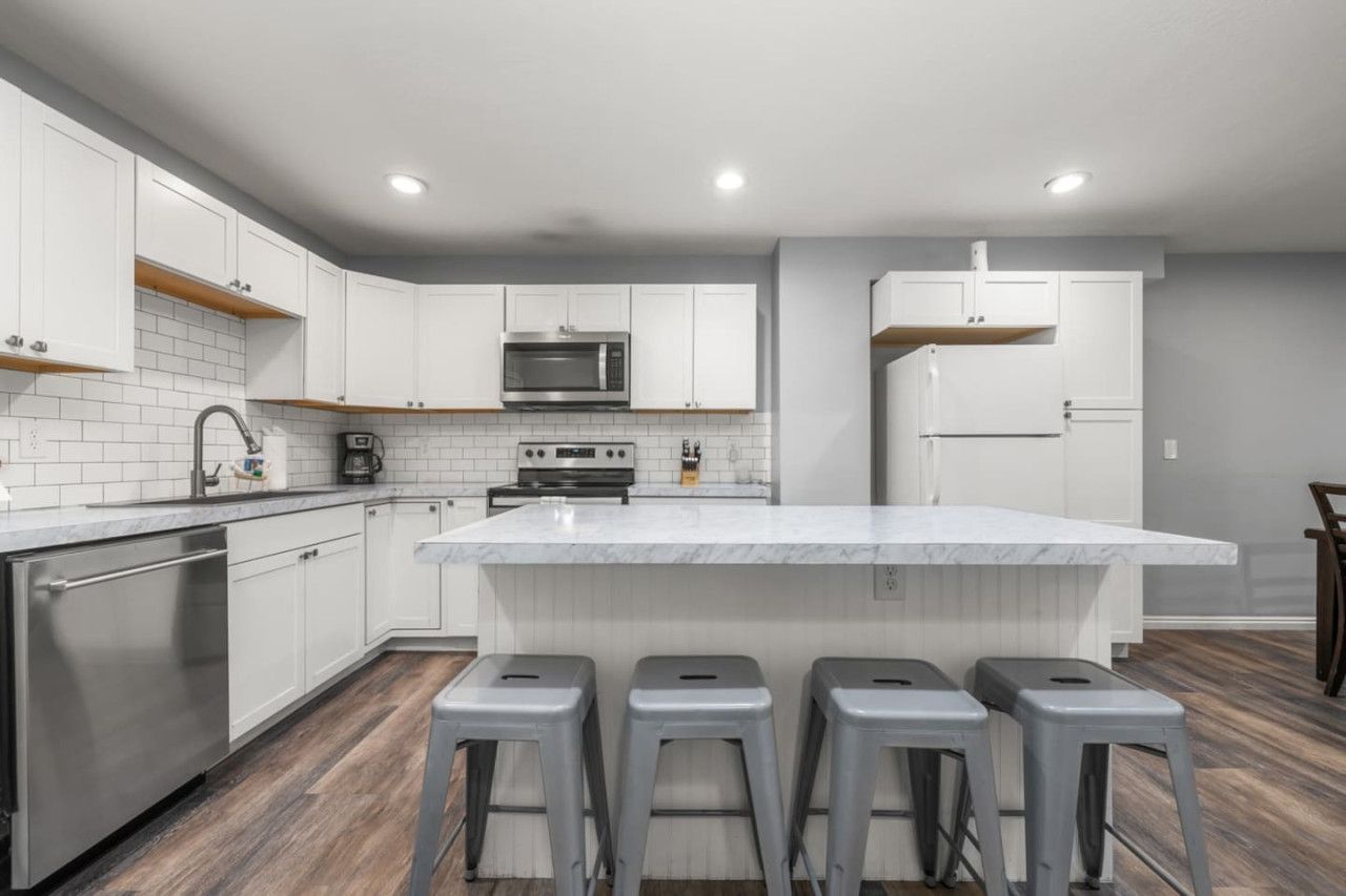 A modern kitchen with white cabinets, a large island with bar stools, stainless steel appliances, and wooden flooring.