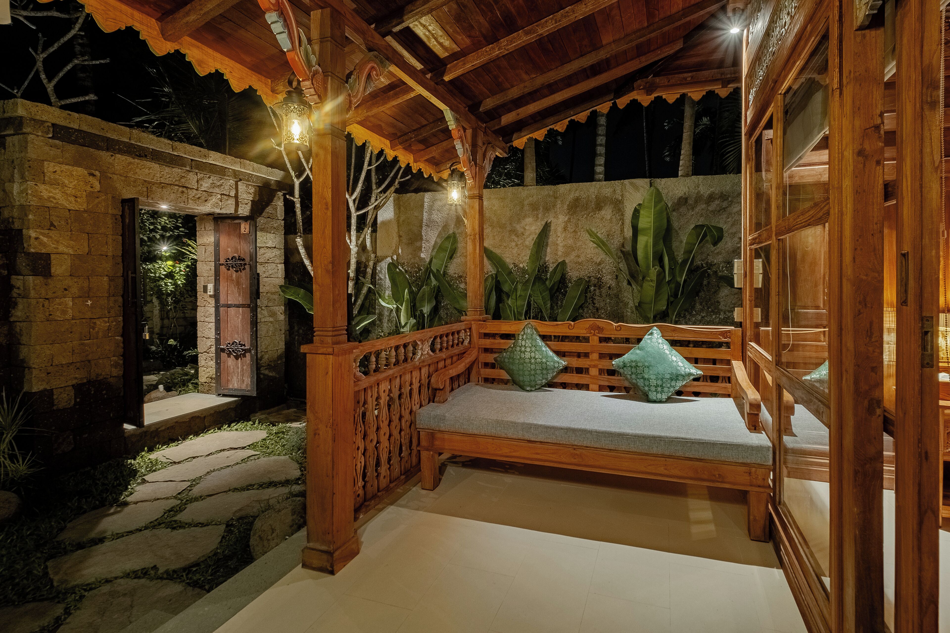 Un banco de madera con cojines en un espacio al aire libre cubierto, con escalones de piedra y plantas tropicales.