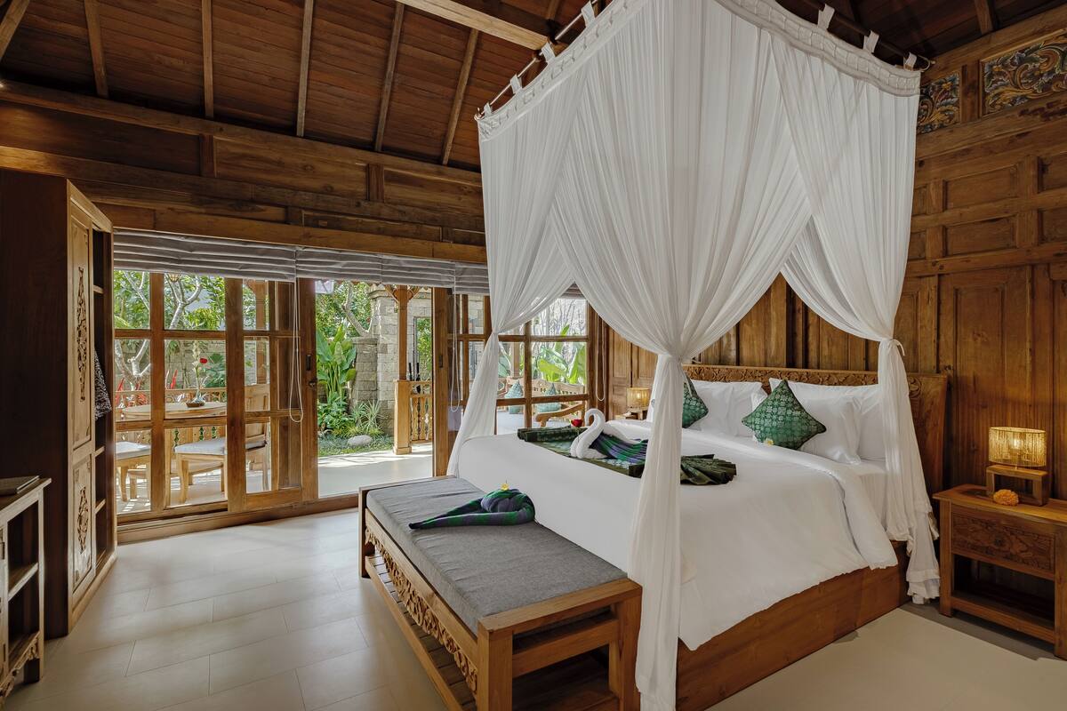A wooden four-poster bed with white bedding, a bench, and a nightstand with a lamp.