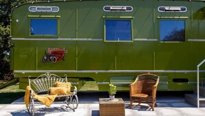 A green RV with a bed, a wicker chair, and a small table with a plant. - Restored Showman's Carriage, King Bed and Pool (Batchmere)
