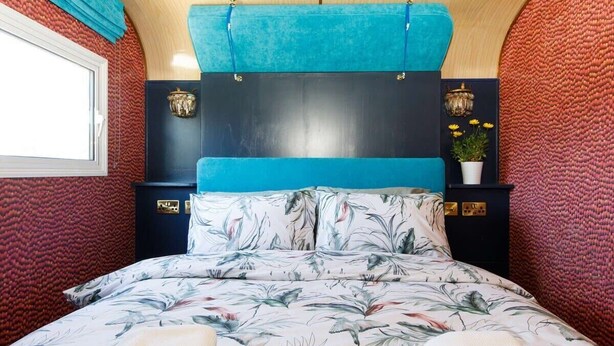 A bedroom with a floral bedspread, a blue headboard, and a decorative wall panel. - Restored Showman's Carriage, King Bed and Pool (Batchmere)