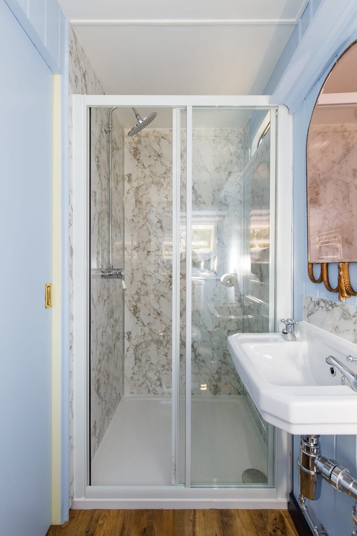 Un baño con cabina de ducha de vidrio, pared de ducha con mosaicos de mármol y lavamanos blanco con grifo dorado.