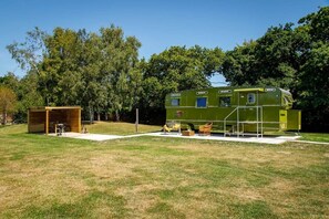 Una autocaravana verde con una estructura pequeña adosada, una banca y zona de barbacoa en un campo cubierto de césped, con árboles al fondo.