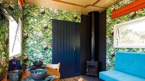 A cozy room with a wooden dining table, a blue sofa, a black stove, and a door with a tropical wallpaper. - Restored Showman's Carriage, King Bed and Pool (Batchmere)