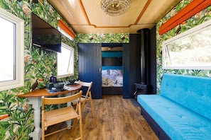 Habitación compacta con suelo de madera, sofá azul, mesa de comedor con sillas, televisor montado en la pared y ventana con cortinas.