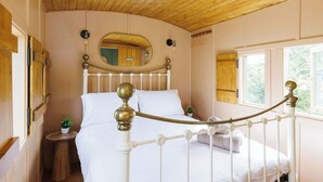 A bedroom with a wooden ceiling, a white bed with a metal headboard, a small bedside table with a plant, and a window with a view of greenery. - Beautifully Restored WW2 Carriage - Sleeps 6 (Batchmere)