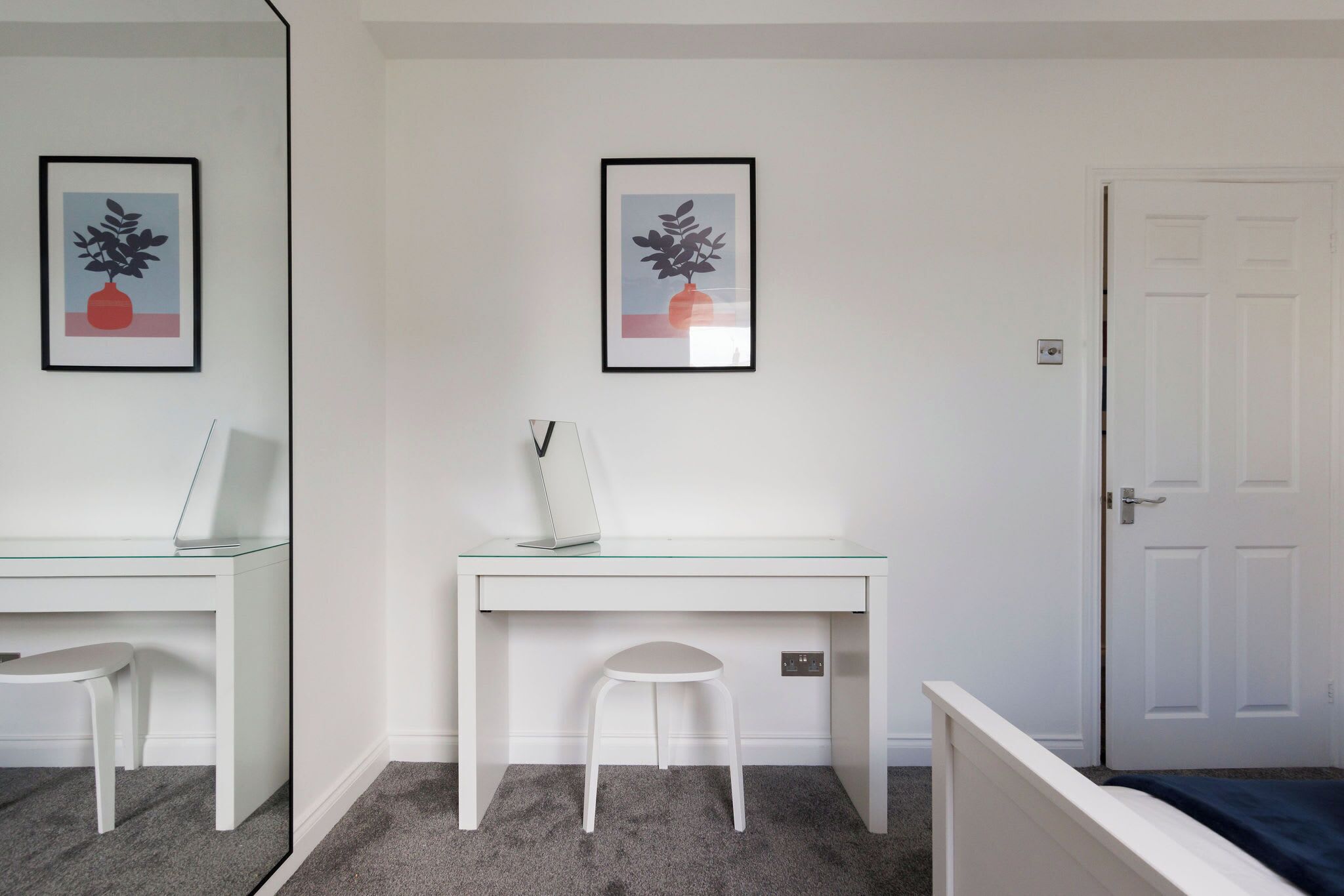 A room with a white desk, a white chair, a small stool, and a door. There are two framed pictures on the wall.