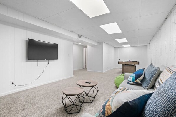 A modern, well-lit room with a flat-screen TV mounted on the wall, two round tables, a sofa with patterned cushions, and a ping pong table.