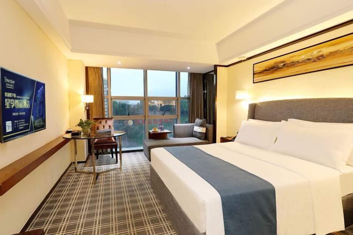 A hotel room with a large bed, a desk with a chair, a small table, and a sofa. There is a window with a view of trees and a building.