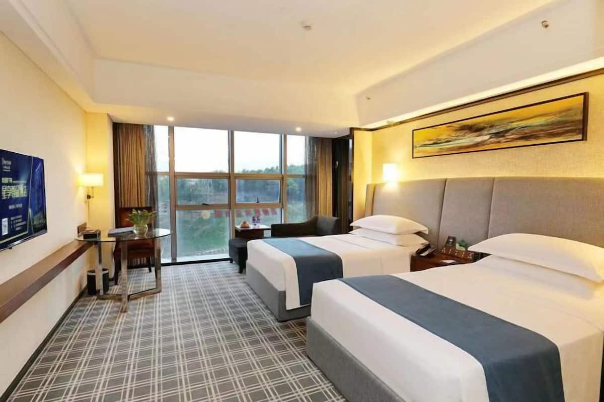 A hotel room with two beds, a TV, a desk with a lamp, and a large window offering a view.
