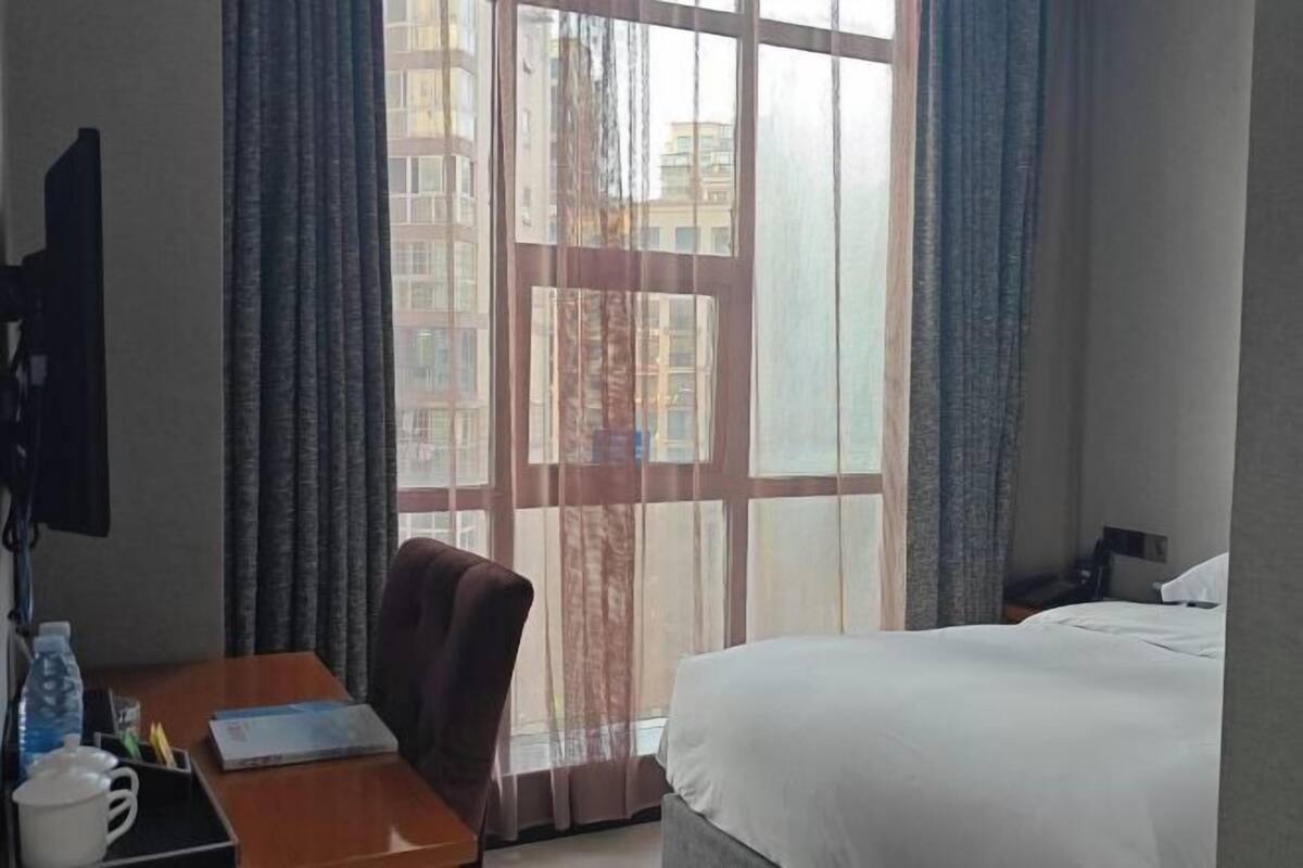 A hotel room with a bed, a desk with a chair, a window with curtains, and a view of buildings outside.