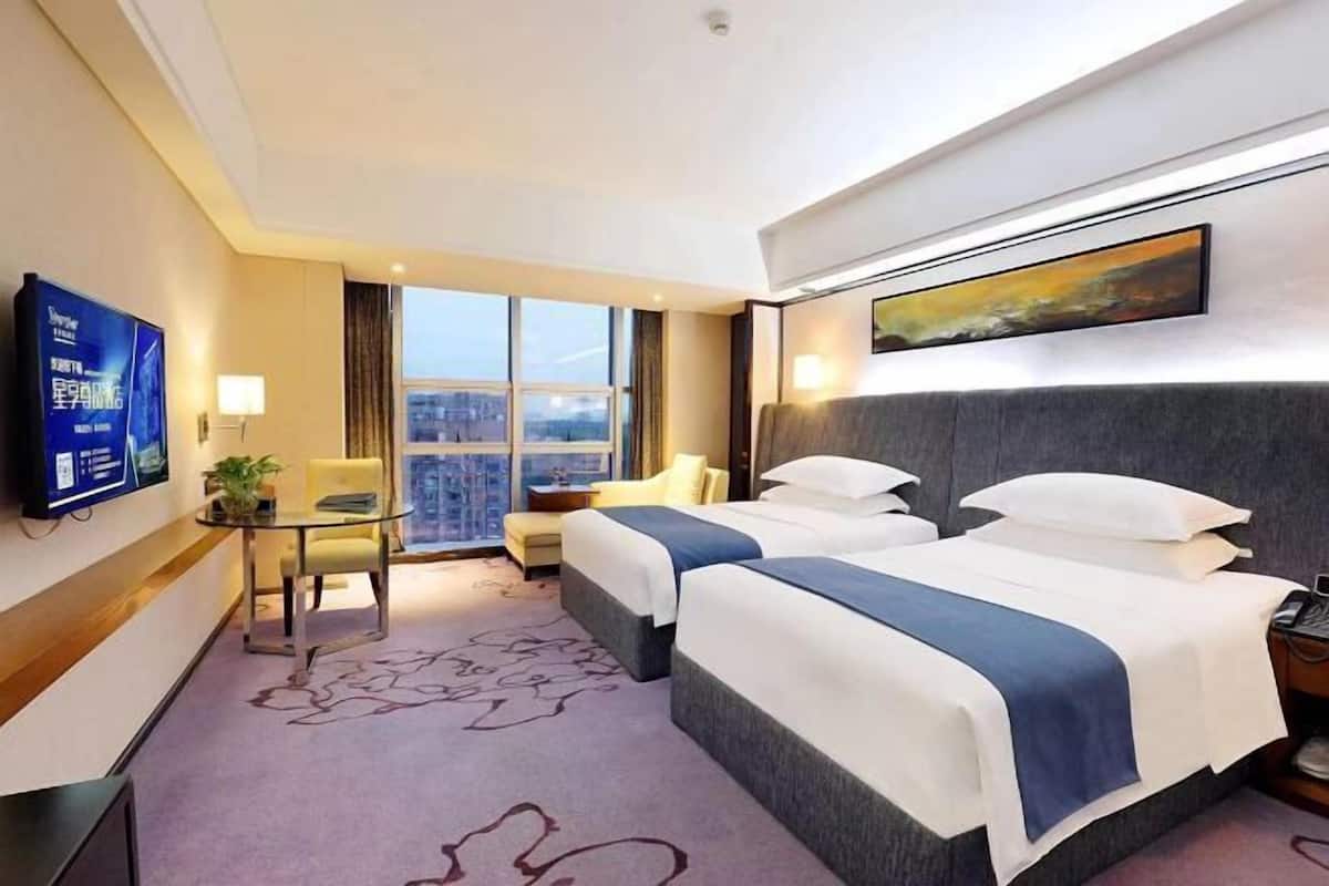 A hotel room with two beds, a TV, a desk, a chair, a window with a city view, and a painting on the wall.