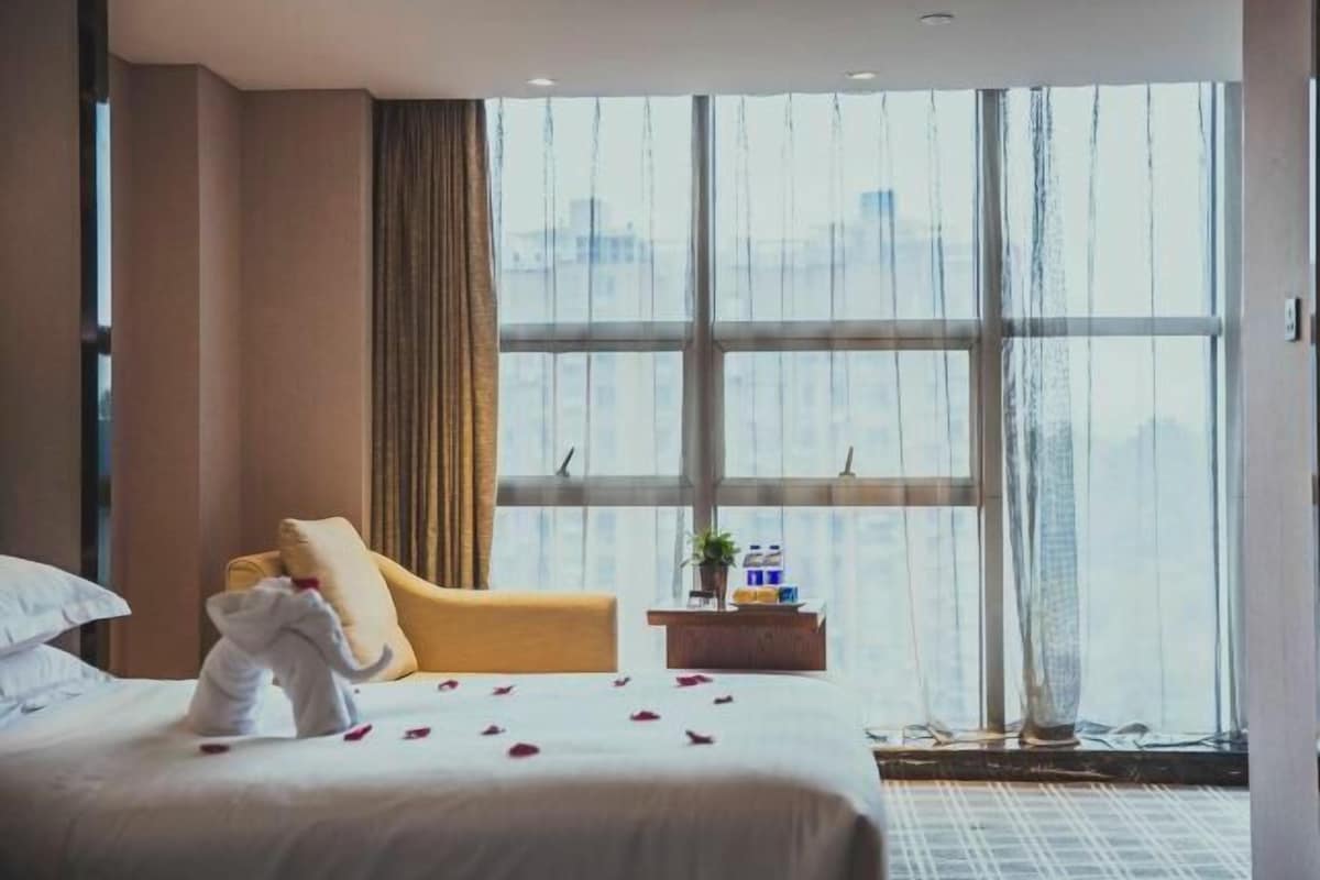 A hotel room with a large window, a bed with white linens, a yellow armchair, and a small table with a plant.