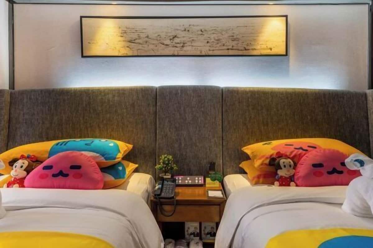 A hotel room with two beds, each with colorful pillows and stuffed toys, a small table with a phone and a plant, and a framed artwork above the beds.