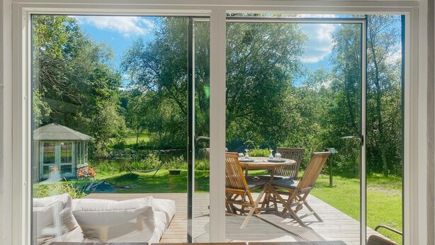 Ein Zimmer mit einer Glasschiebetür, die auf eine Außenterrasse mit Tisch, Stühlen, einer Hängematte und einem kleinen Garten mit Teich führt.