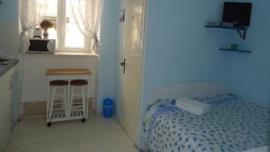 A small, compact room with a bed, a desk with two chairs, a window with curtains, a ceiling fan, a mounted TV, and a trash bin. - Dubrovnik Cozy central Studio with street View (Dubrovnik)