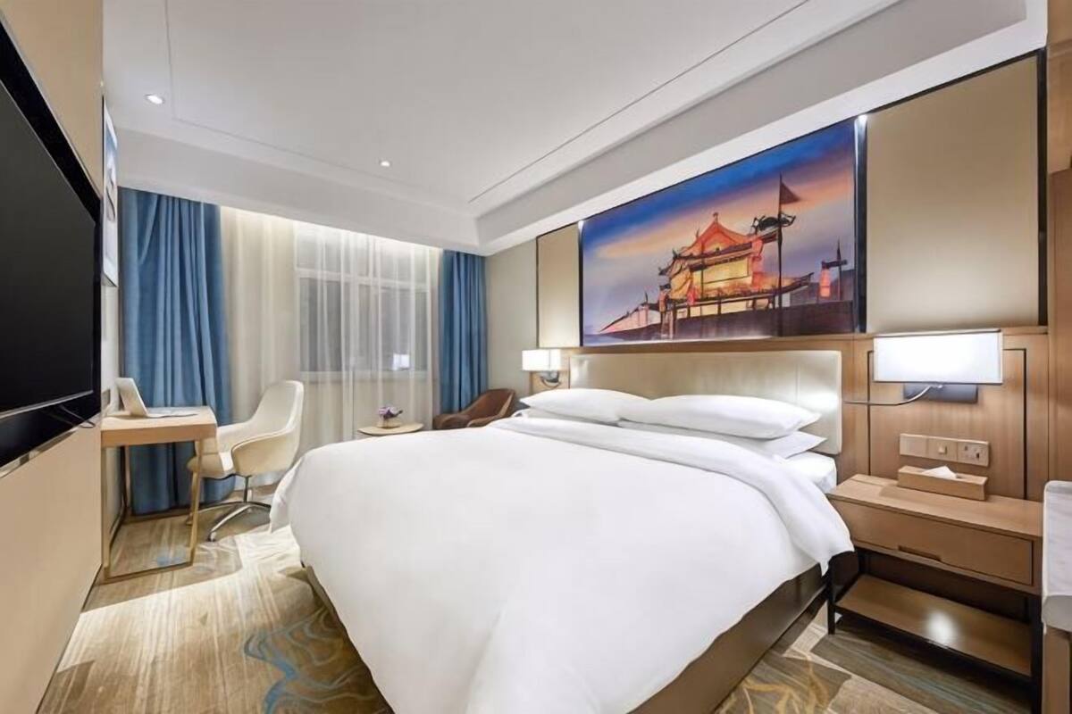 A hotel room with a large bed, a desk, a chair, a TV, and a painting of a building.