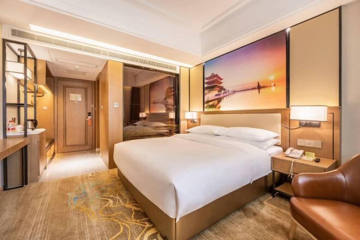 A hotel room with a bed, bedside table, lamp, and a painting of a bridge.