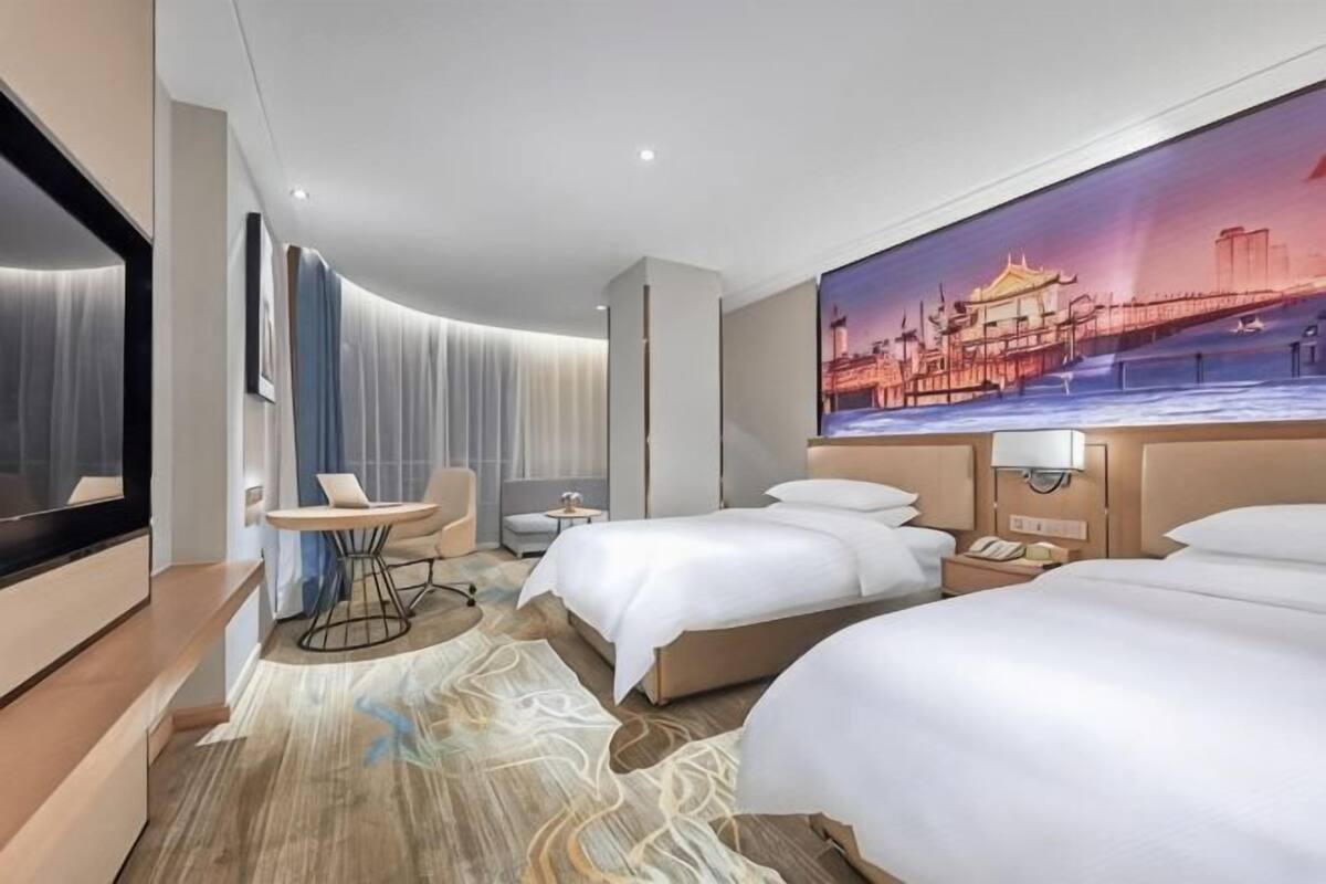 A hotel room with two beds, a desk, a chair, a TV, and a large cityscape painting on the wall.