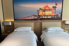 Habitación de hotel con dos camas, una mesita de noche, una lámpara y un cuadro grande en la pared que muestra un edificio tradicional.