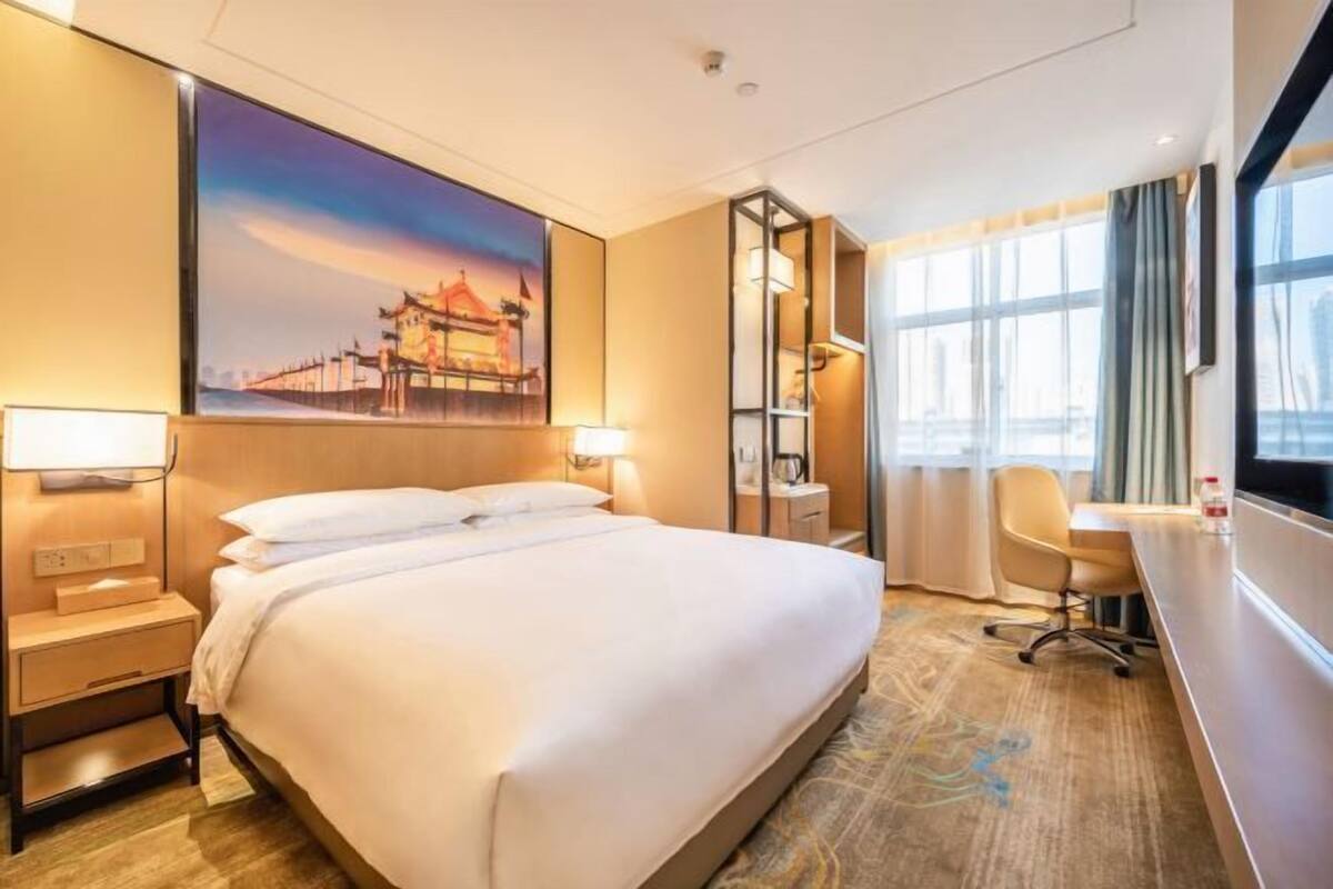 A hotel room with a bed, bedside tables, a desk, a chair, a large window, and a painting on the wall.