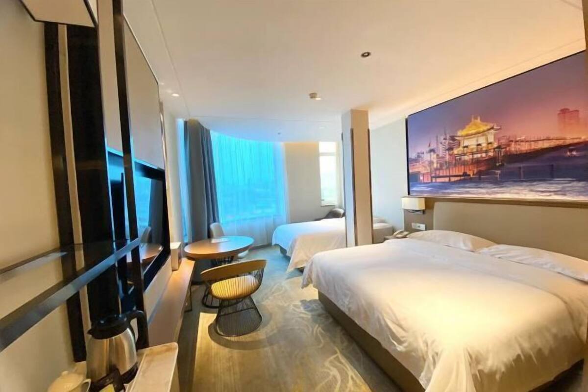 A hotel room with a bed, a desk, a chair, a window with curtains, and a painting on the wall.