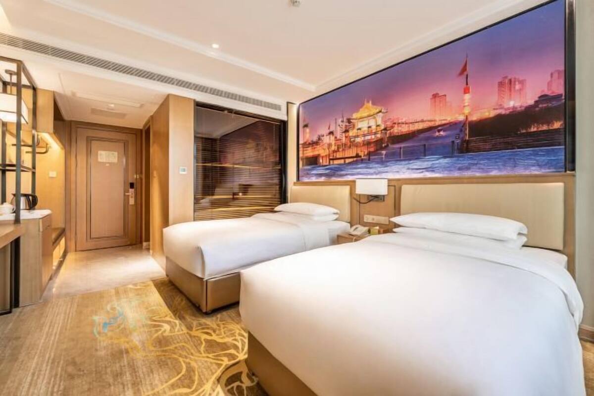 A hotel room with two beds, a large window with a cityscape view, a desk with a lamp, and a door leading to a bathroom.