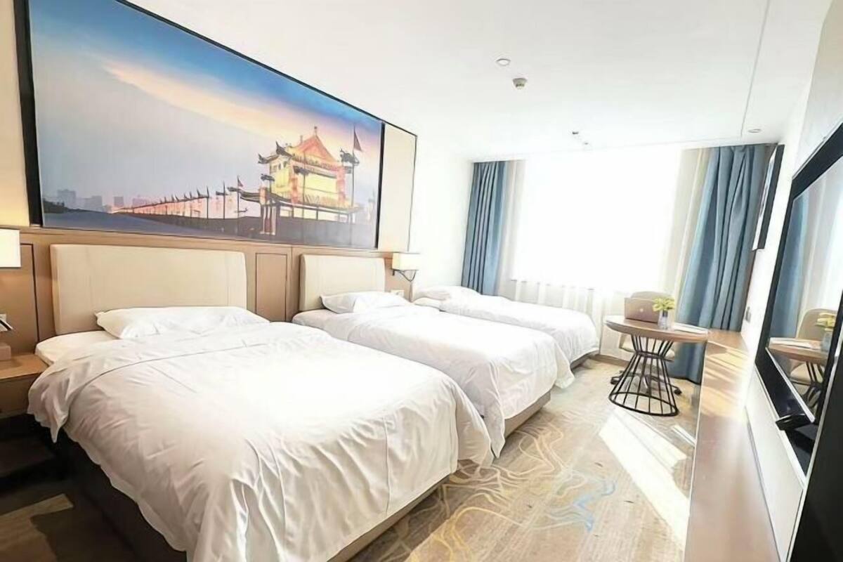 A hotel room with two beds, a large painting of a building, a small table with a chair, and a window with curtains.
